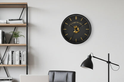 Black wall clock with gold accents and Bitcoin symbol, placed on a white wall with a plant and small pot.
