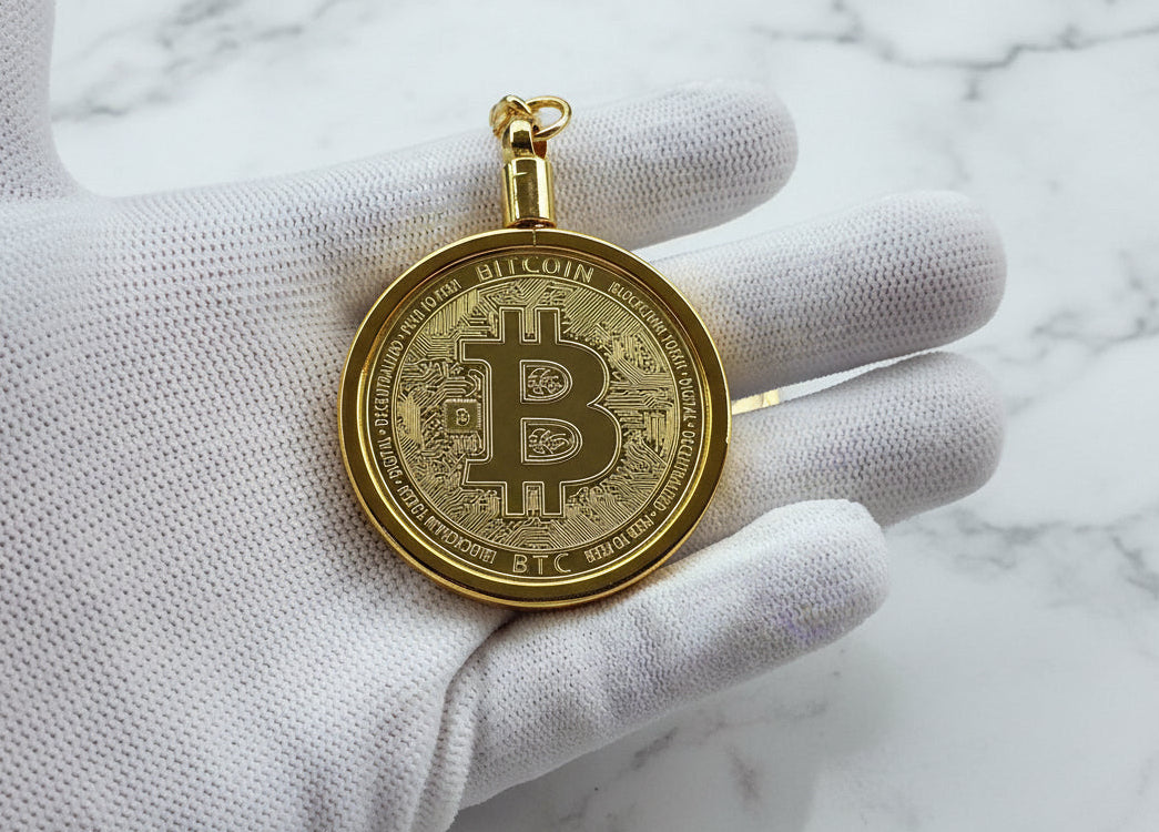 Gold Bitcoin pendant held by a gloved hand against a marble background